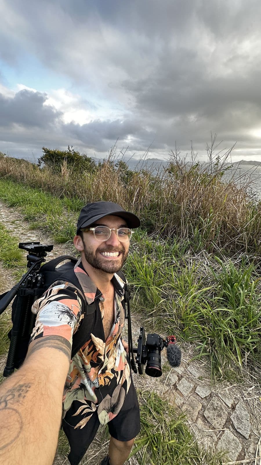 Selfie of Nando in a cap and floral shirt, with tripod and camera, on a seaside trail