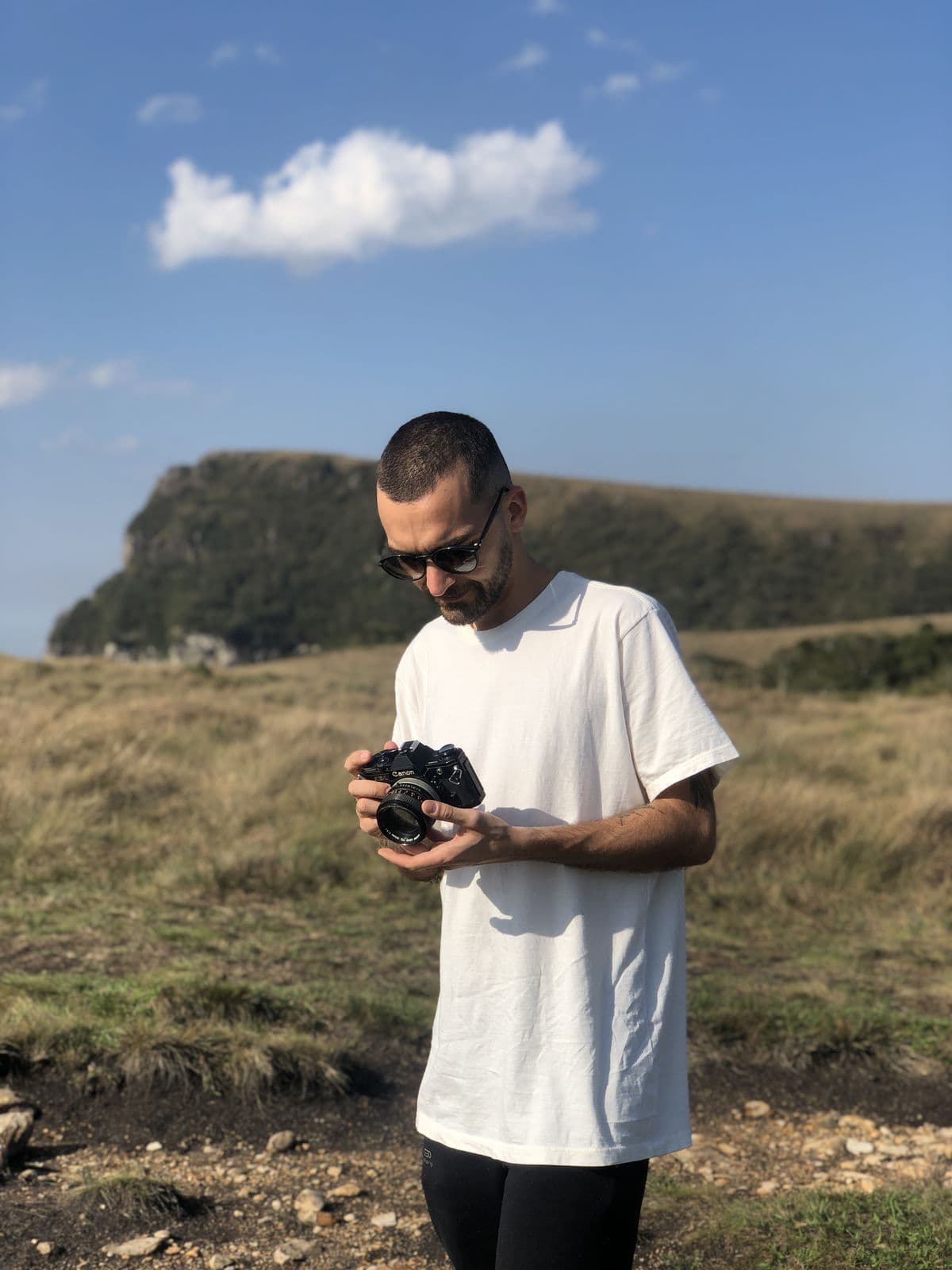 Nando in a white t-shirt looking at the camera in his hands, in a field with a rock wall behind