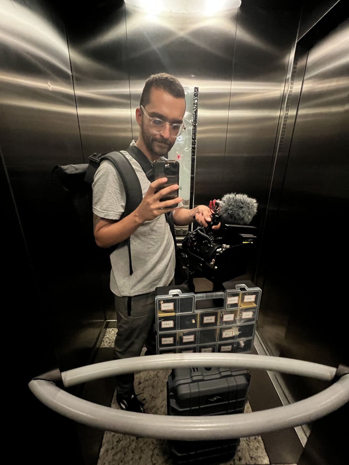 Nando in an elevator mirror, with a backpack, a camera and an equipment case