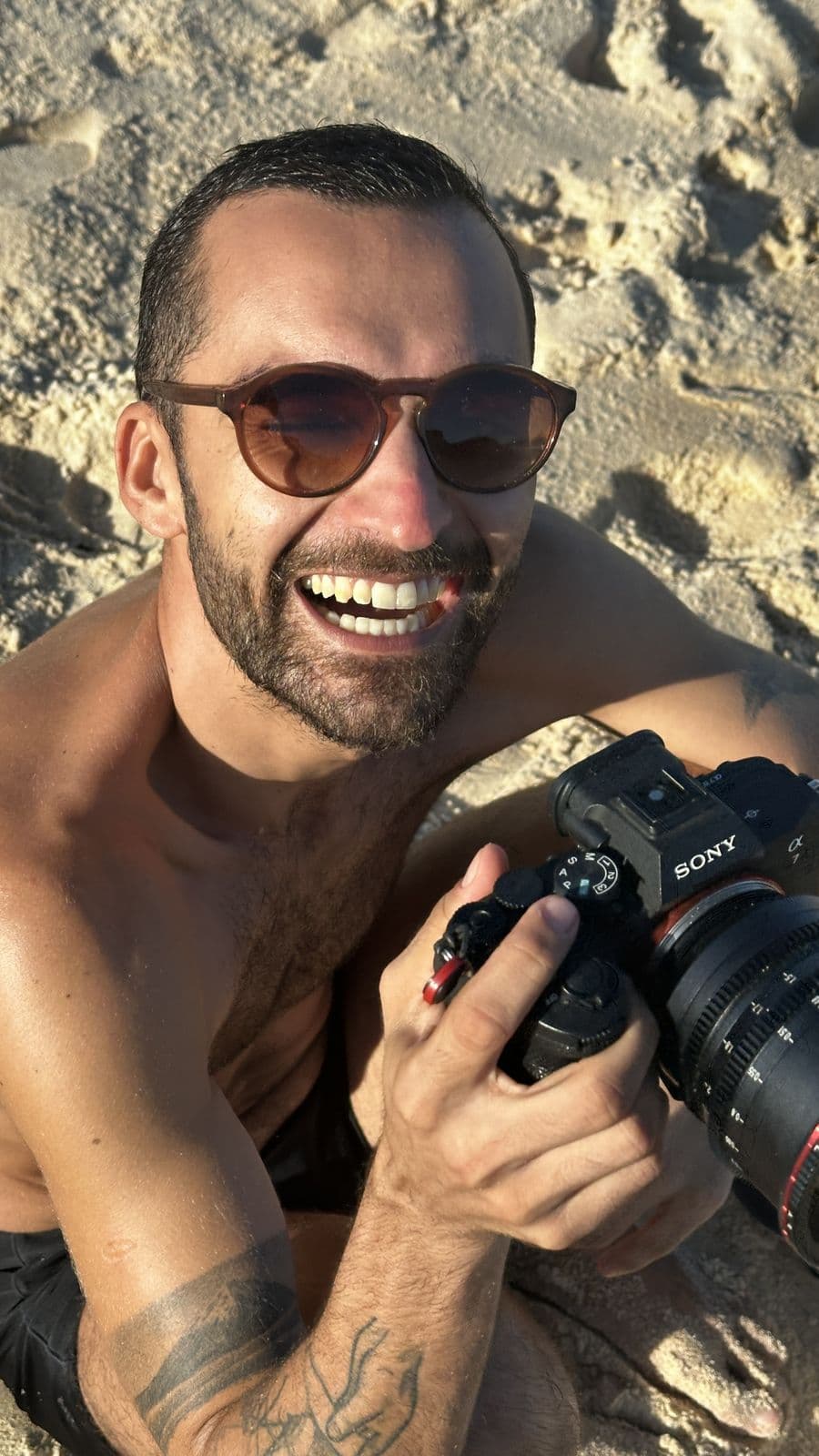 Nando smiling on the sand, shirtless, holding a camera