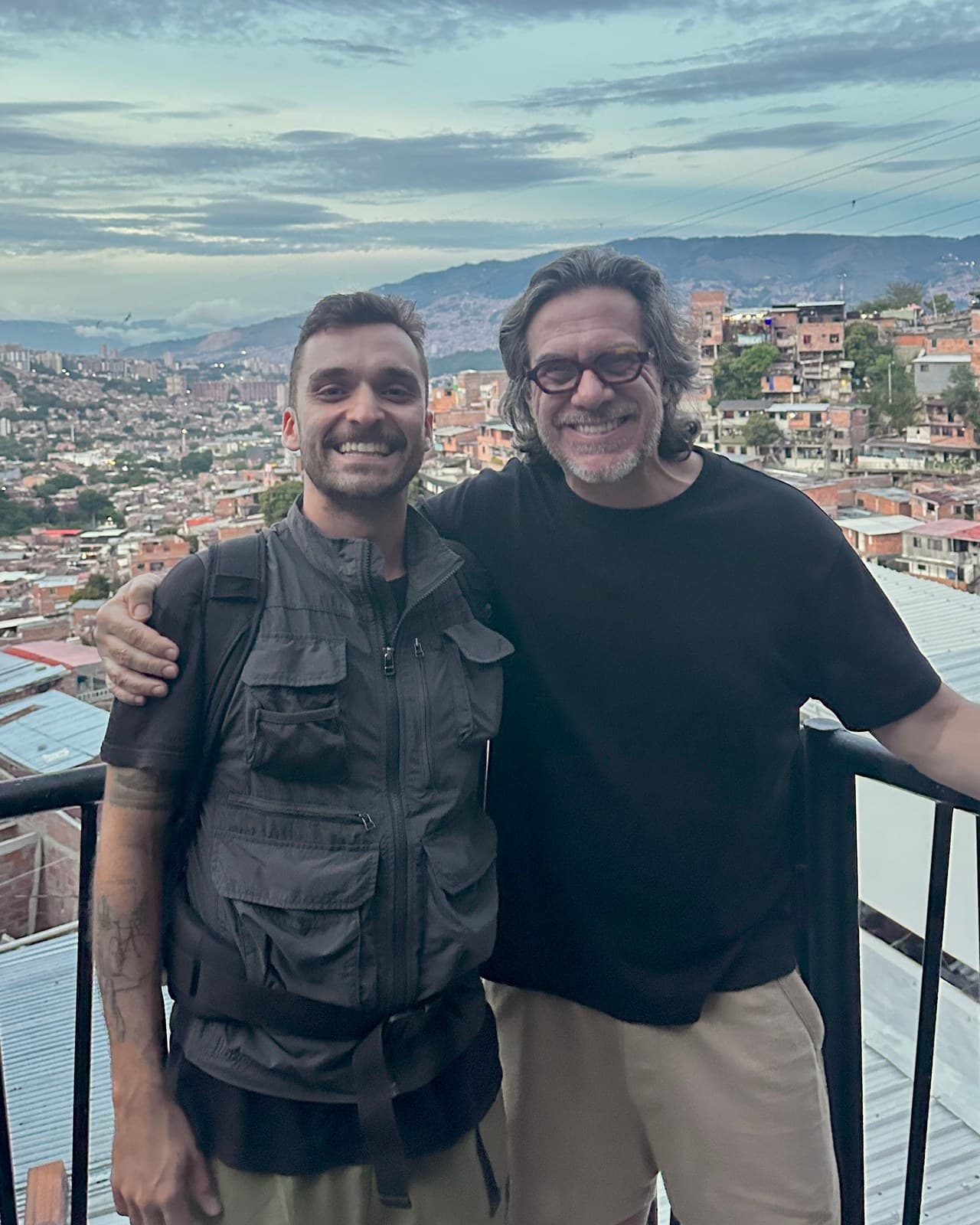 Nando and César Paz arm in arm at a lookout, with the hills of Medellín behind them