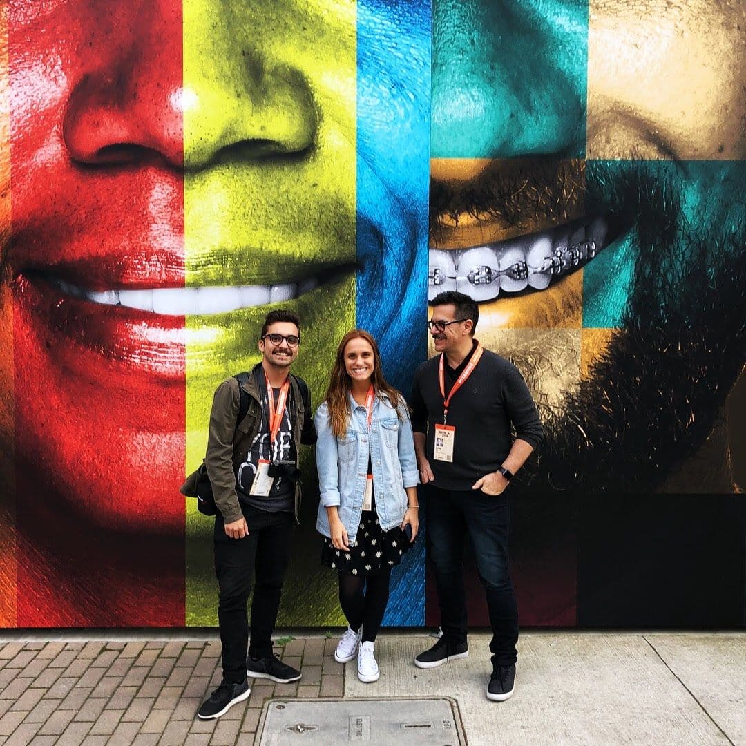 Three people with festival badges in front of a colorful mural of smiling faces