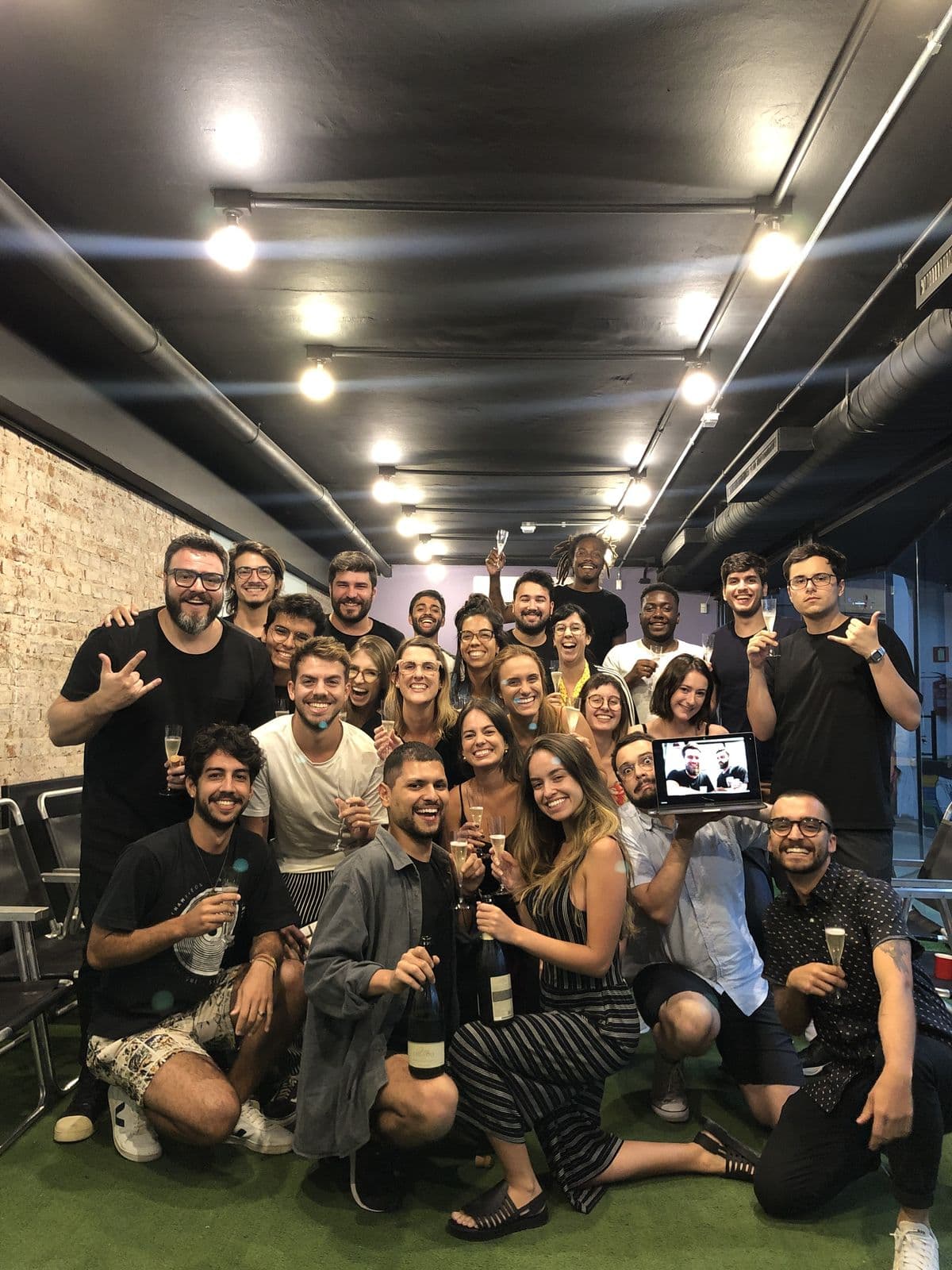 The team gathered with glasses raised, in an office with a dark ceiling and hanging bulbs