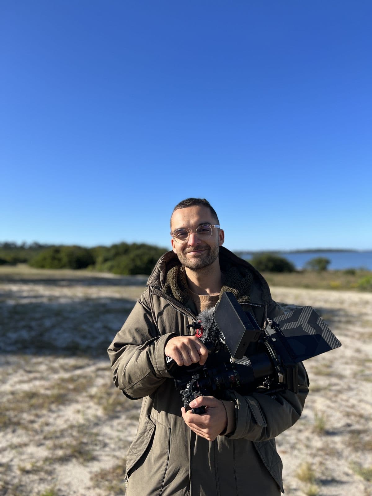 Nando in a coat holding a camera, in a field of sand and grass under a blue sky