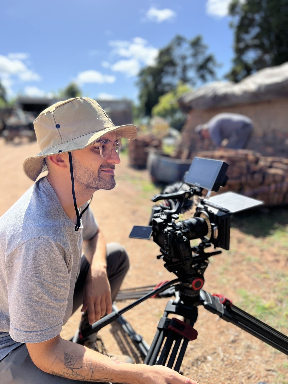Operator in a hat next to a camera on a tripod, with bricks and a brickworks behind