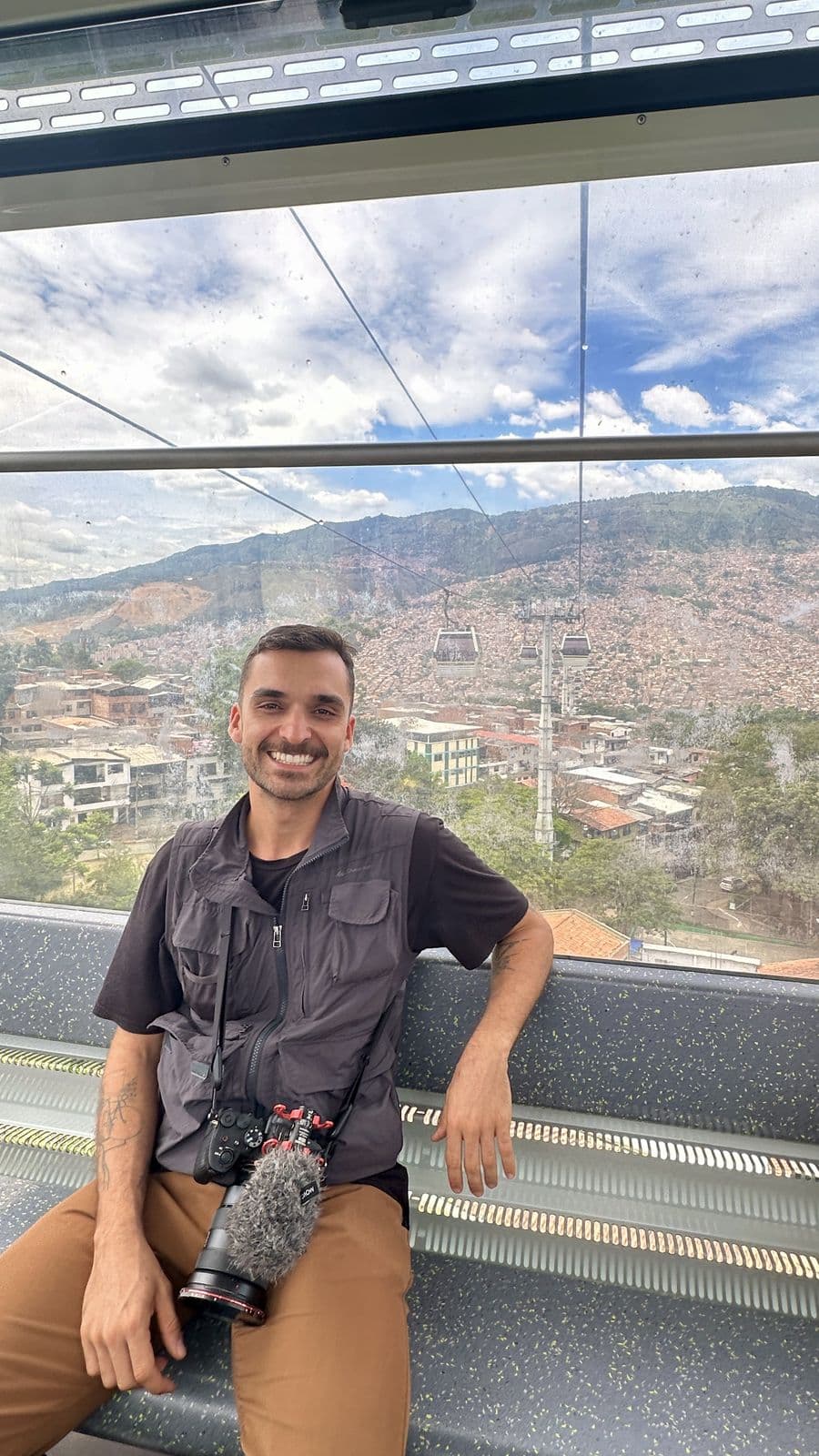 Nando sitting in a cable car cabin, camera on his lap and the city behind