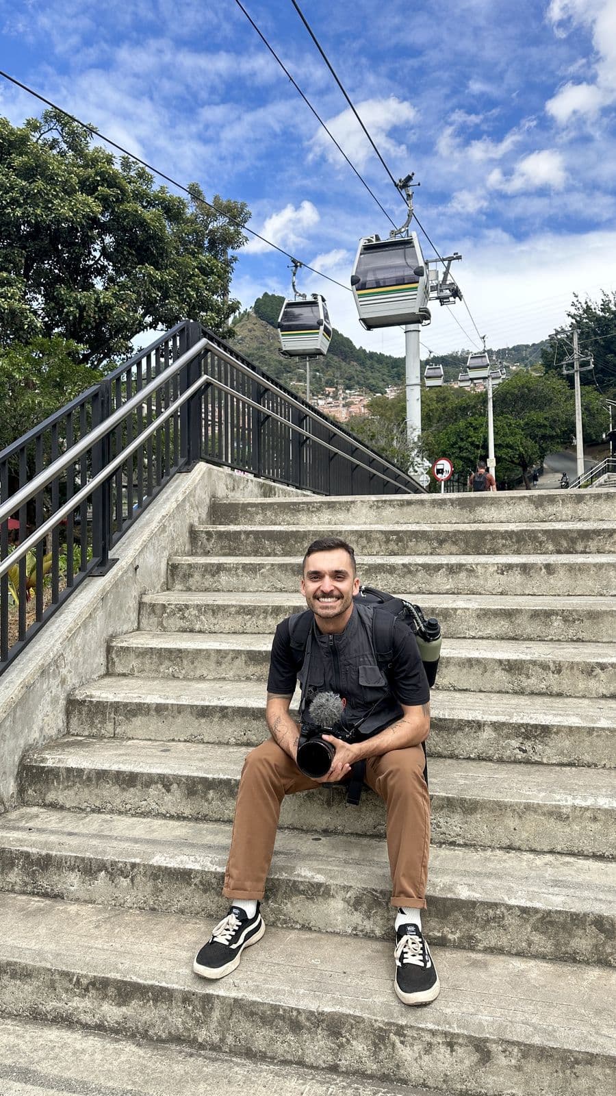 Nando sitting on concrete steps, camera in hand, with the cable car passing behind