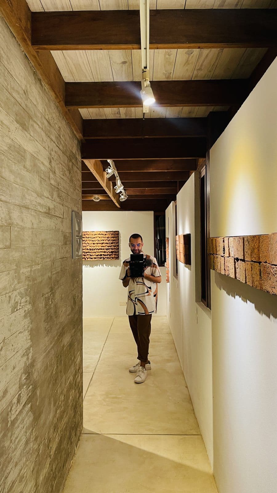 Narrow gallery corridor with brick pieces on the walls and Nando filming at the far end