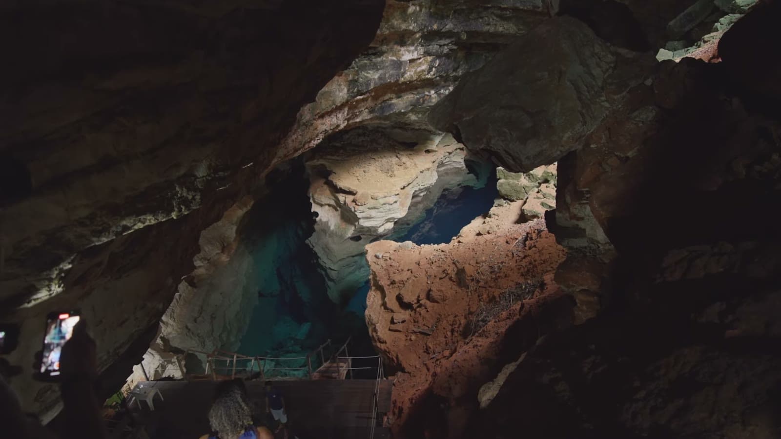 Interior da caverna visto do alto da escada, com a água azul-turquesa do poço lá embaixo entre as rochas