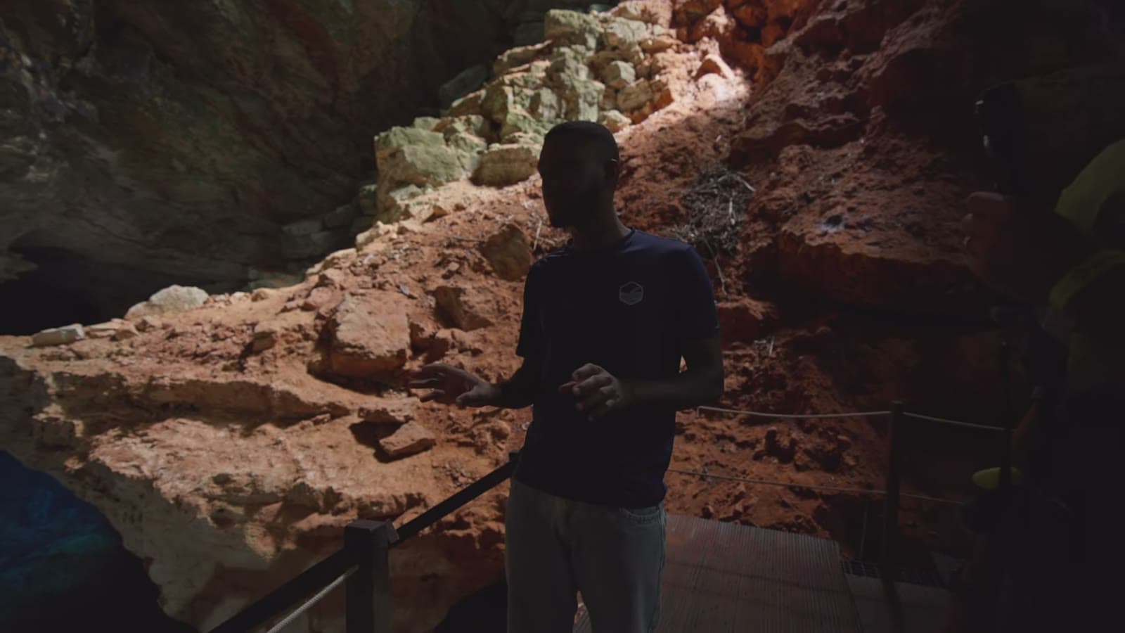 O guia do parque em pé na plataforma de acesso, explicando as regras, com as rochas avermelhadas da caverna atrás