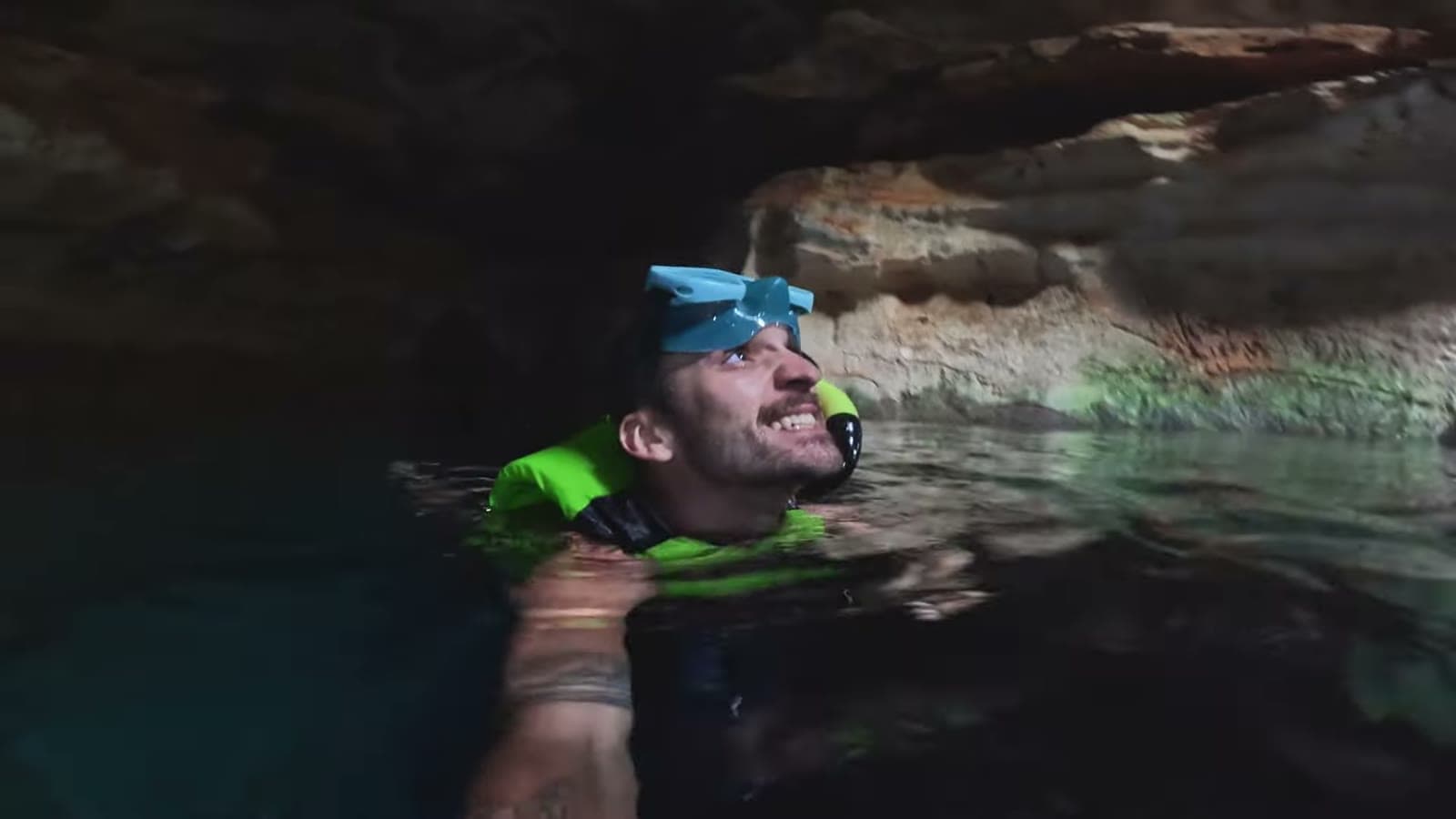 Nando flutuando de colete e máscara de mergulho, sorrindo, sob o teto de pedra da caverna