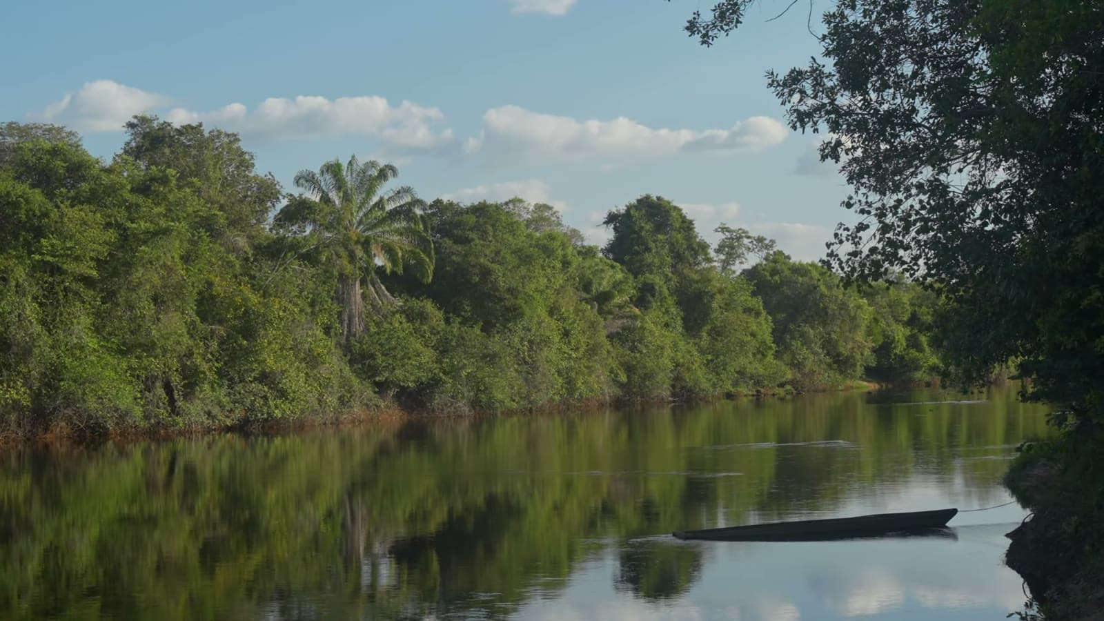Rio de águas calmas refletindo a mata verde da margem e o céu azul com nuvens, com uma canoa parada na água