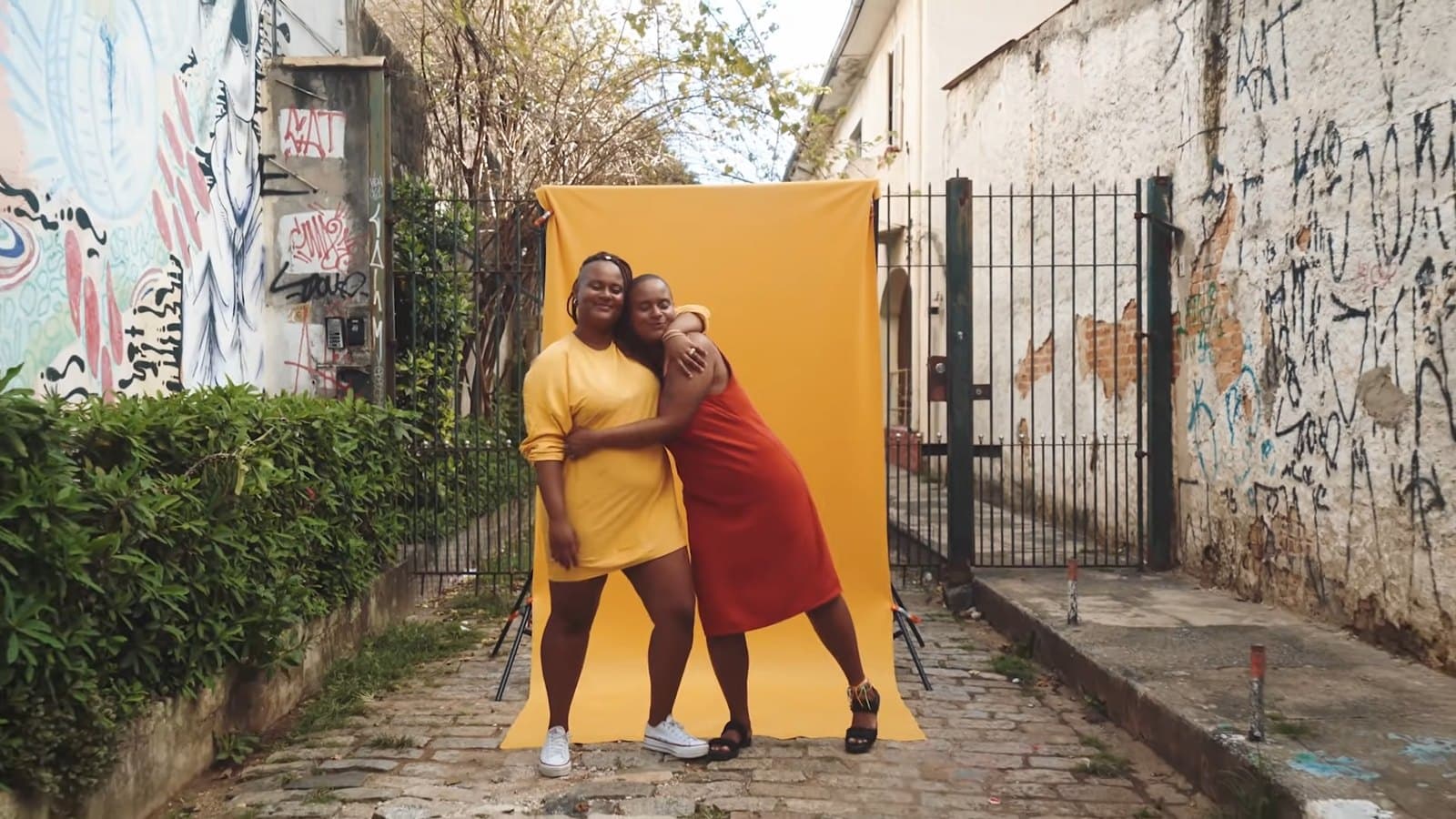 Las dos amigas abrazadas frente a un fondo amarillo montado en un callejón con grafitis