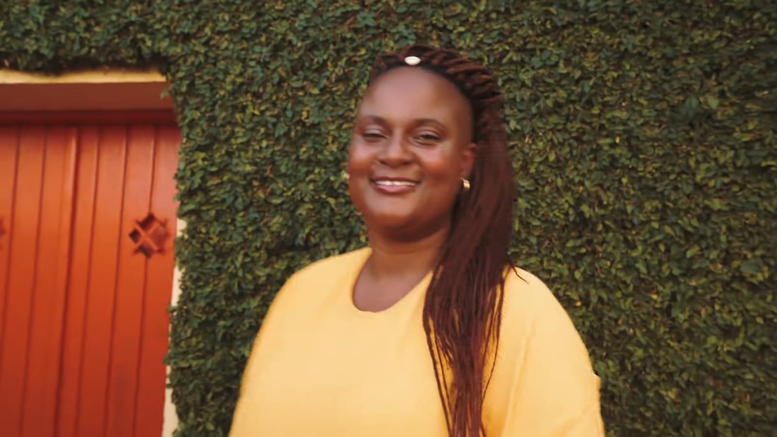 Mujer de amarillo sonríe frente a un muro de hiedra, junto a una puerta naranja
