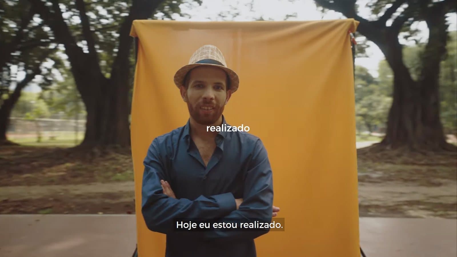 Un hombre con sombrero y brazos cruzados frente a un fondo amarillo, entre árboles