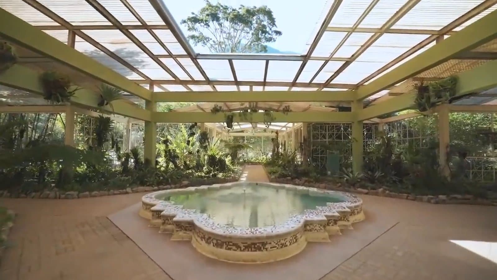 Estufa de teto vazado, cheia de plantas, com um chafariz de mosaico no centro