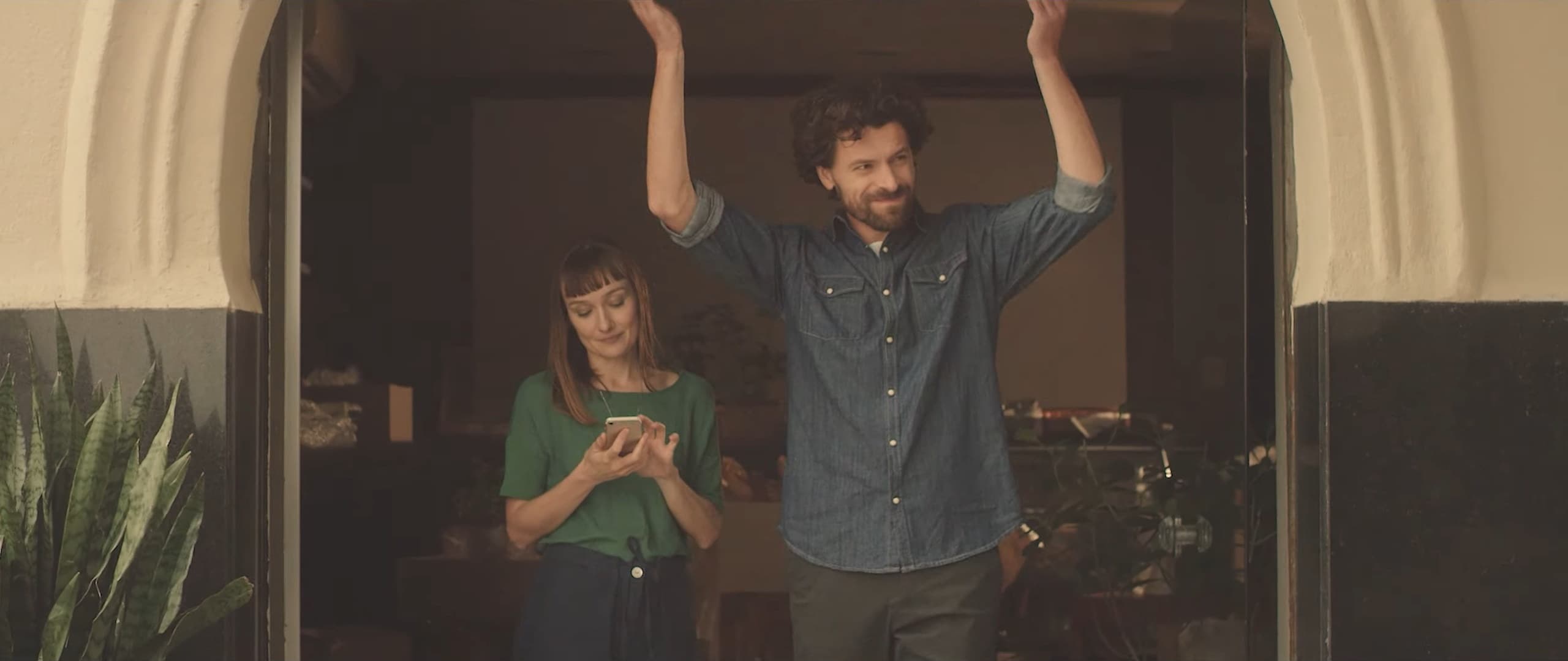A couple at their front door: he dances with his arms raised, she smiles at her phone