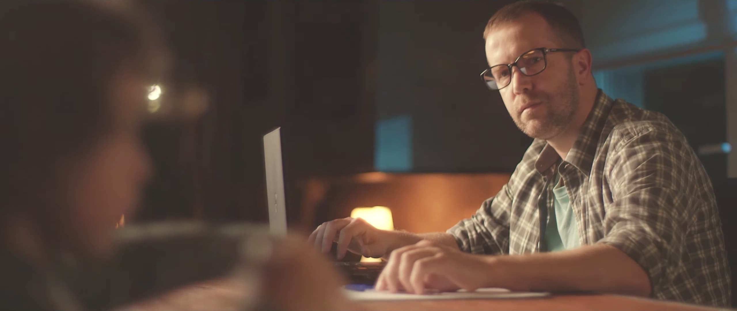A man in glasses at his laptop looks at the camera