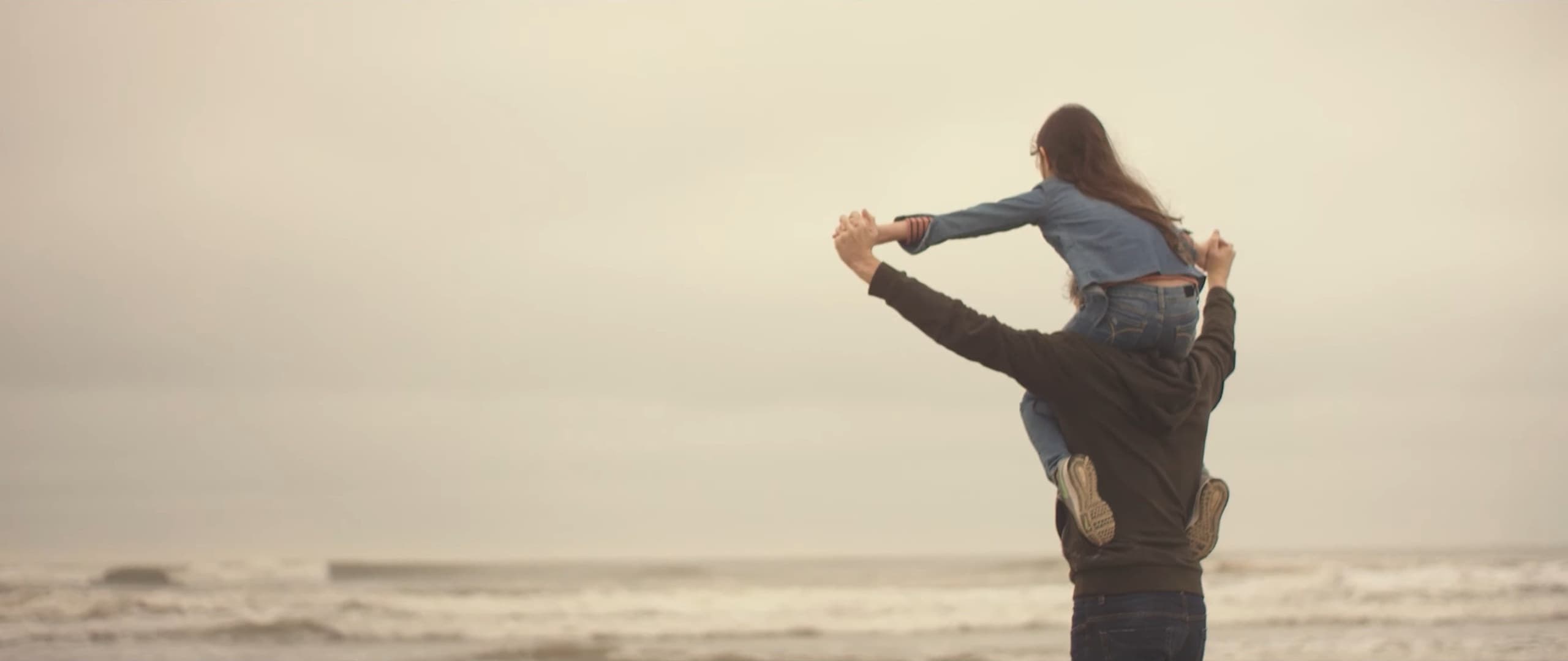 Seen from behind by the sea, a man with his daughter on his shoulders, arms outstretched