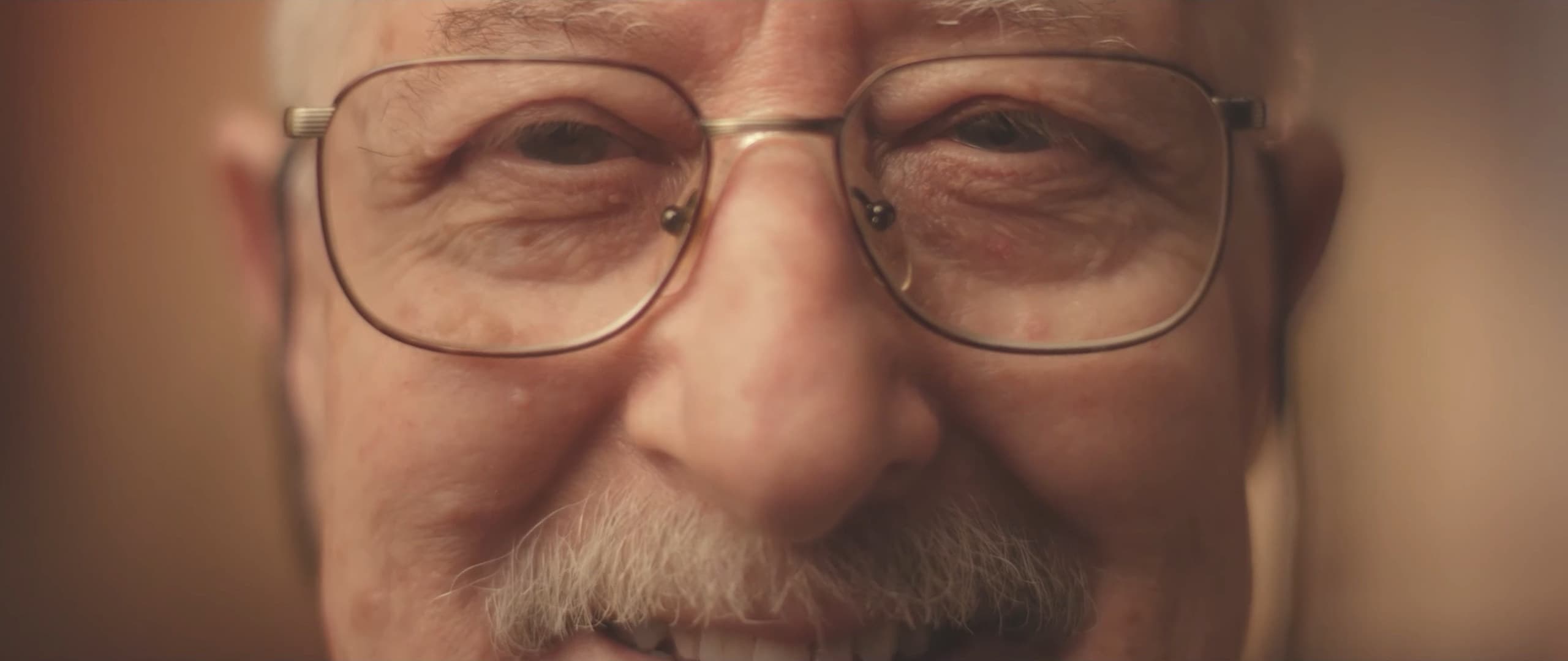 An older man with glasses and a white moustache smiles at the camera