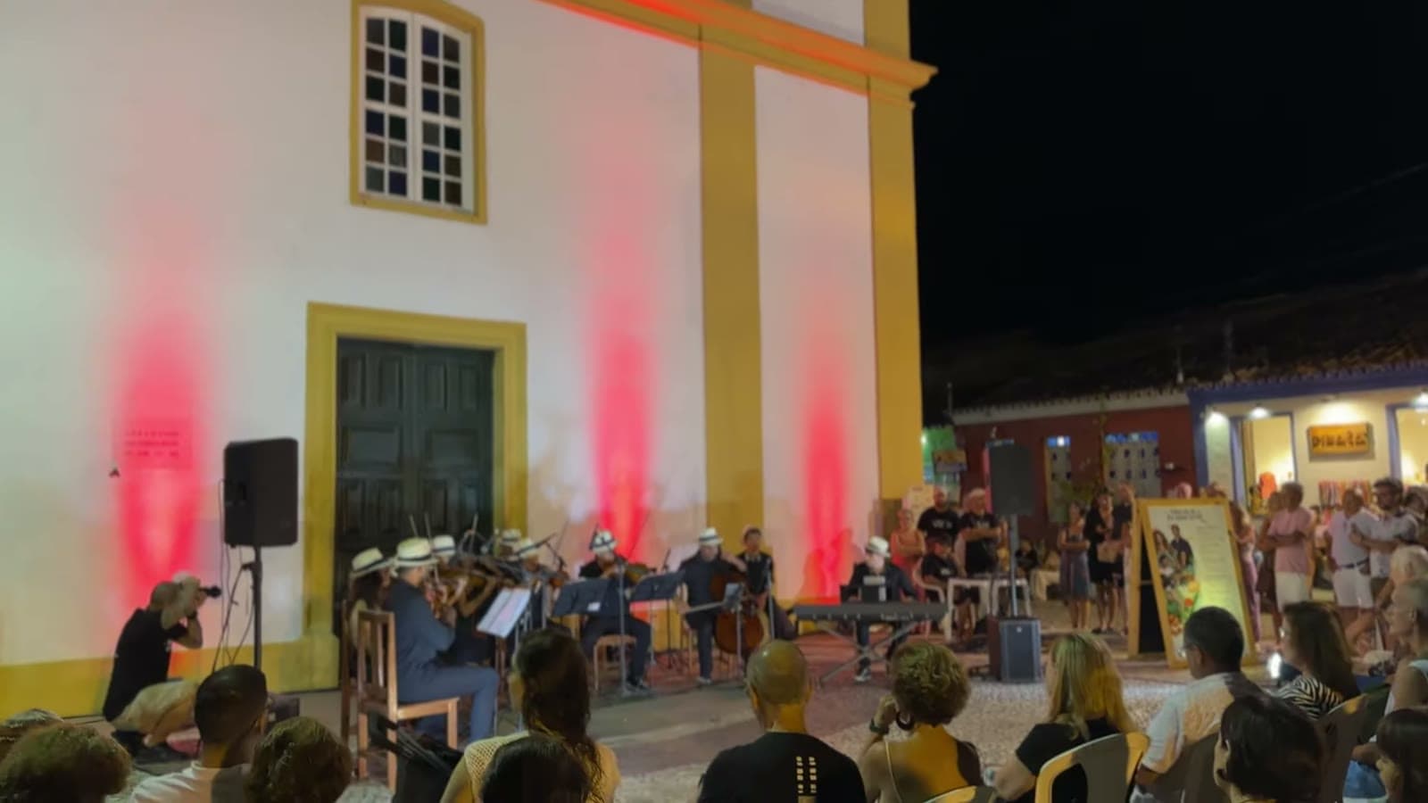 Orchestra musicians playing in front of the church lit up in red, with the audience seated in the square