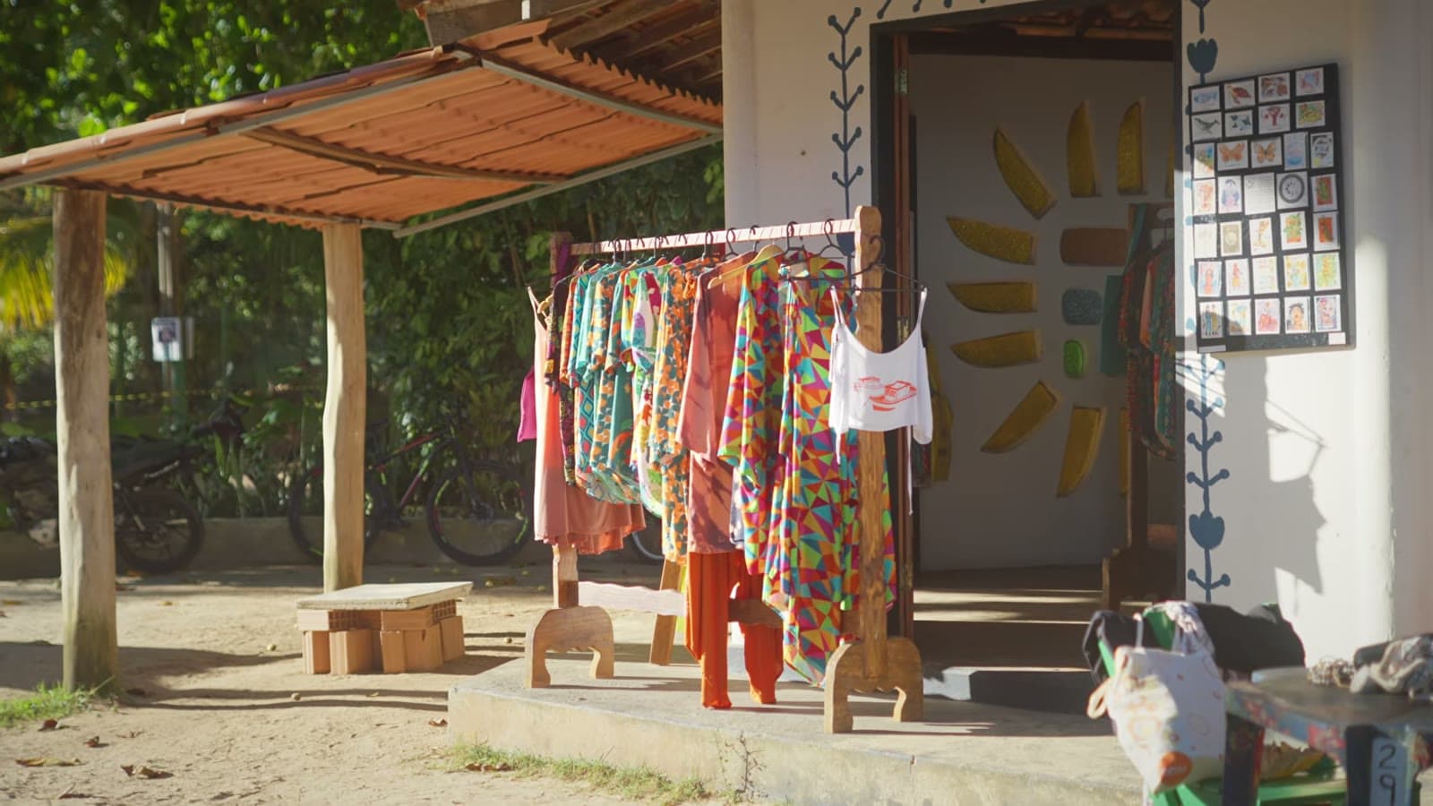 Perchero con ropa colorida de estampados geométricos frente a una tiendita de artesanía, en una calle de arena