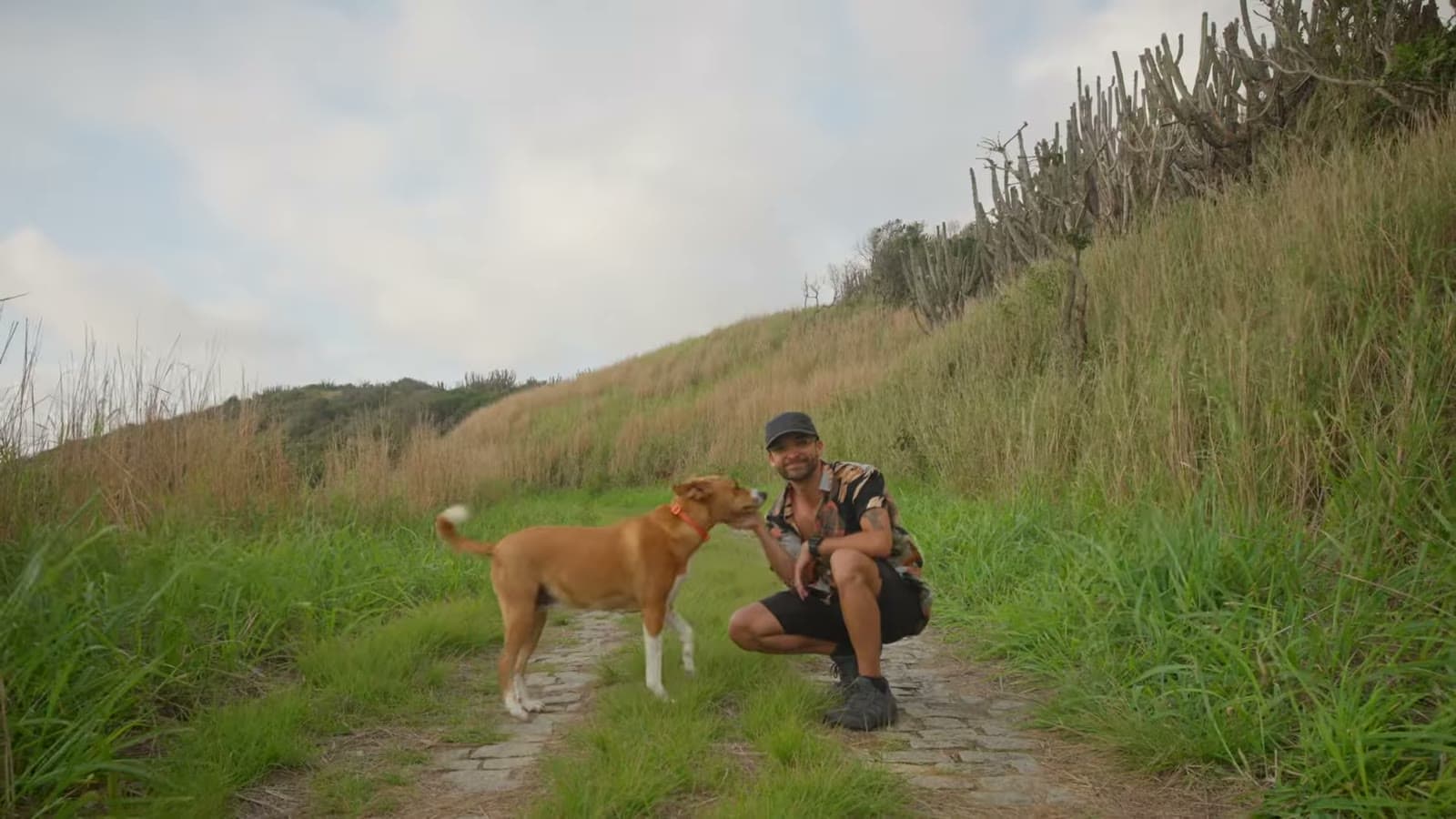 Nando agachado numa estradinha de terra entre o capim, fazendo carinho na Nina