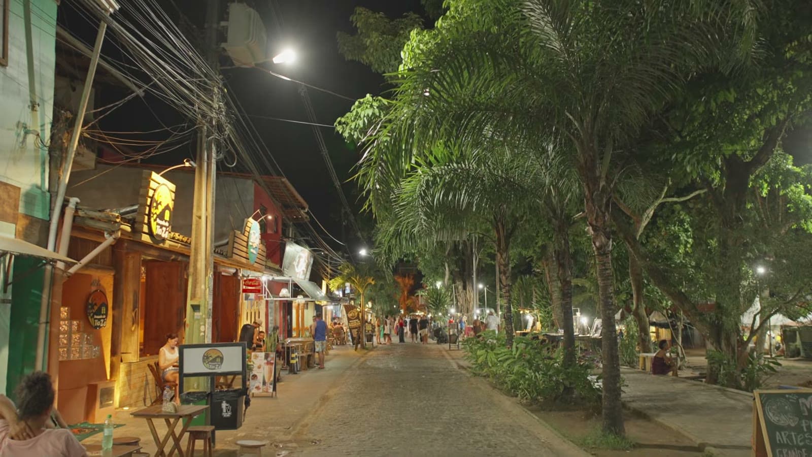 Pituba street at night, with lit-up bars, tables on the sidewalk and trees along the street