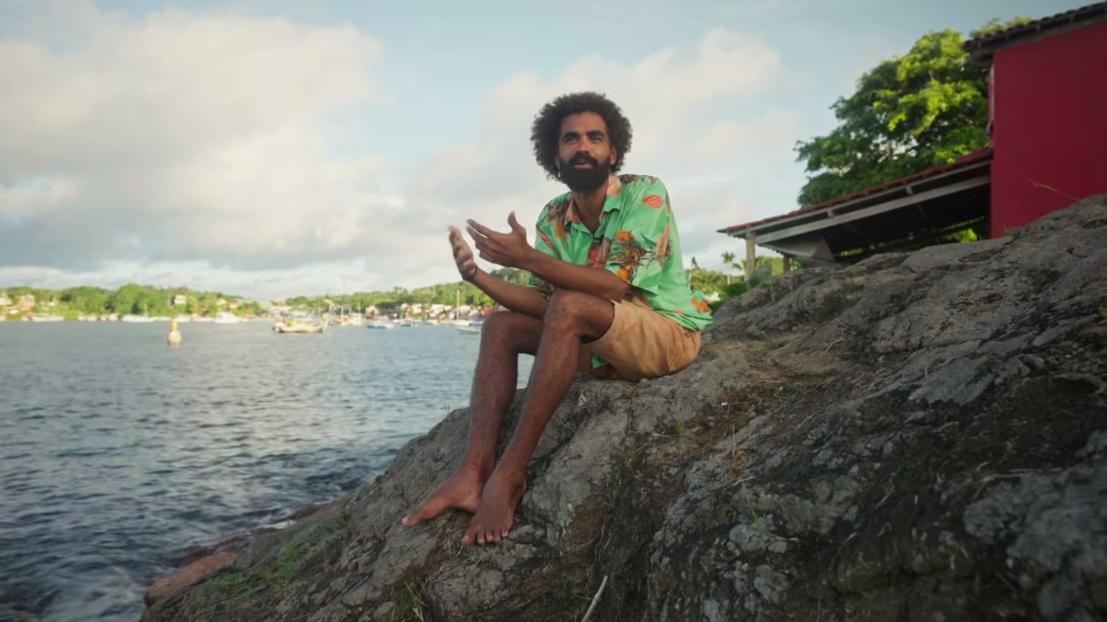 Devid sitting on a rock by the water, talking, with boats and the other shore in the background