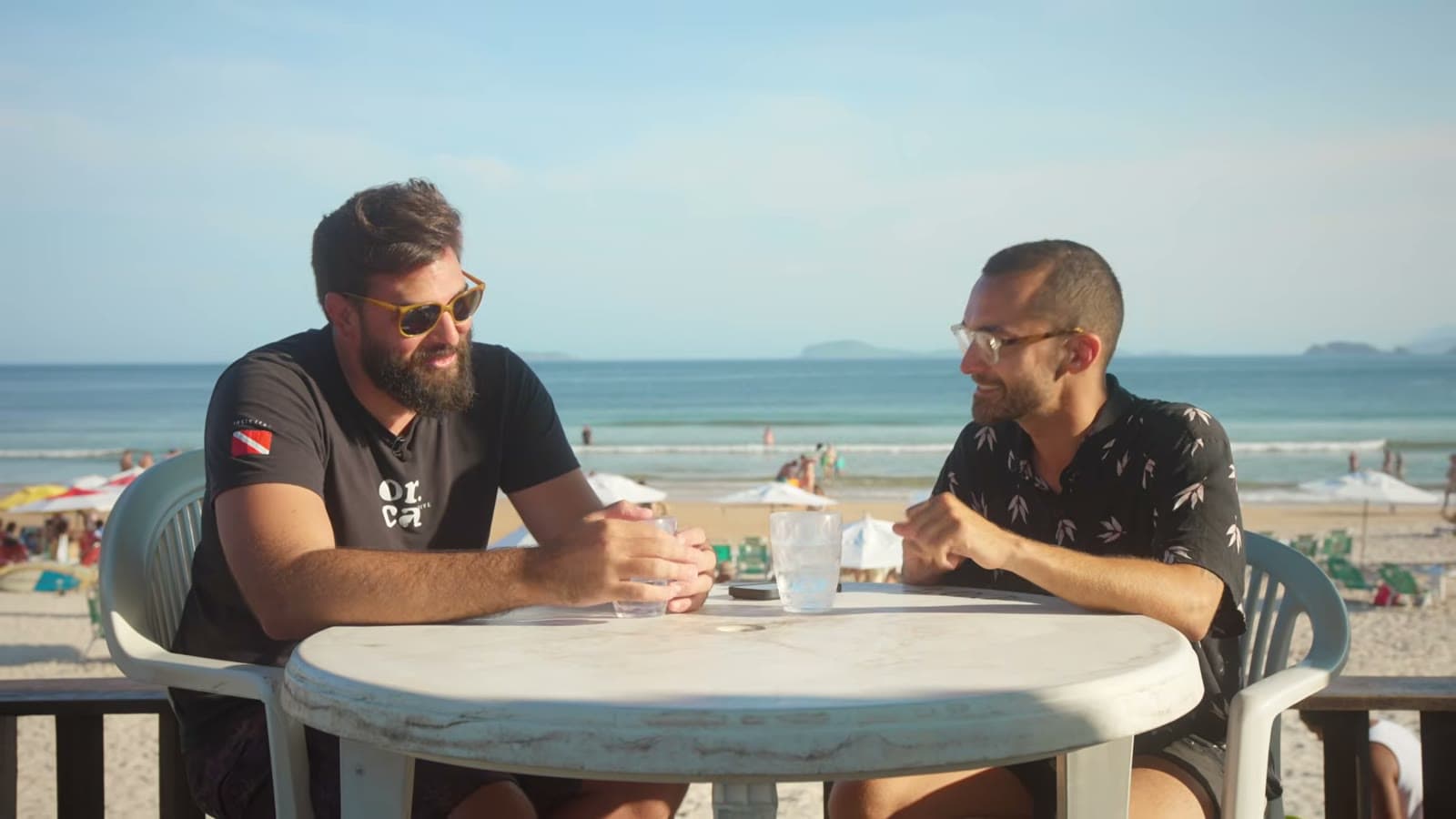 Nicholas, in sunglasses and an Orca Dive t-shirt, talking with Nando at a table facing Praia de Geribá