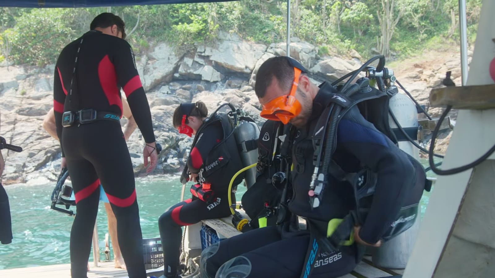 Divers in wetsuits putting on their gear on the boat, near a rocky shore