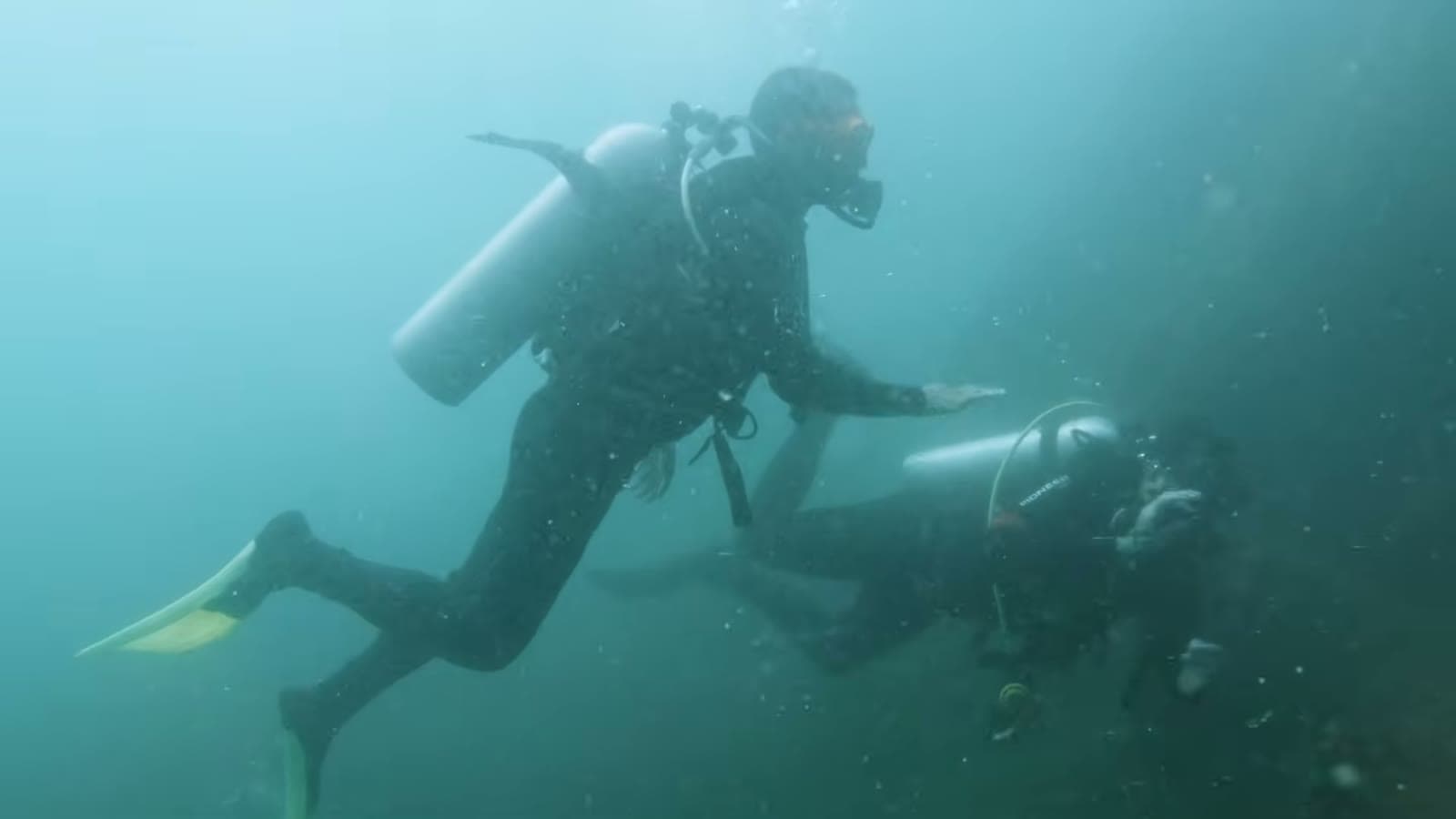 Two divers with tanks on their backs swimming close to the bottom, in blue-green water