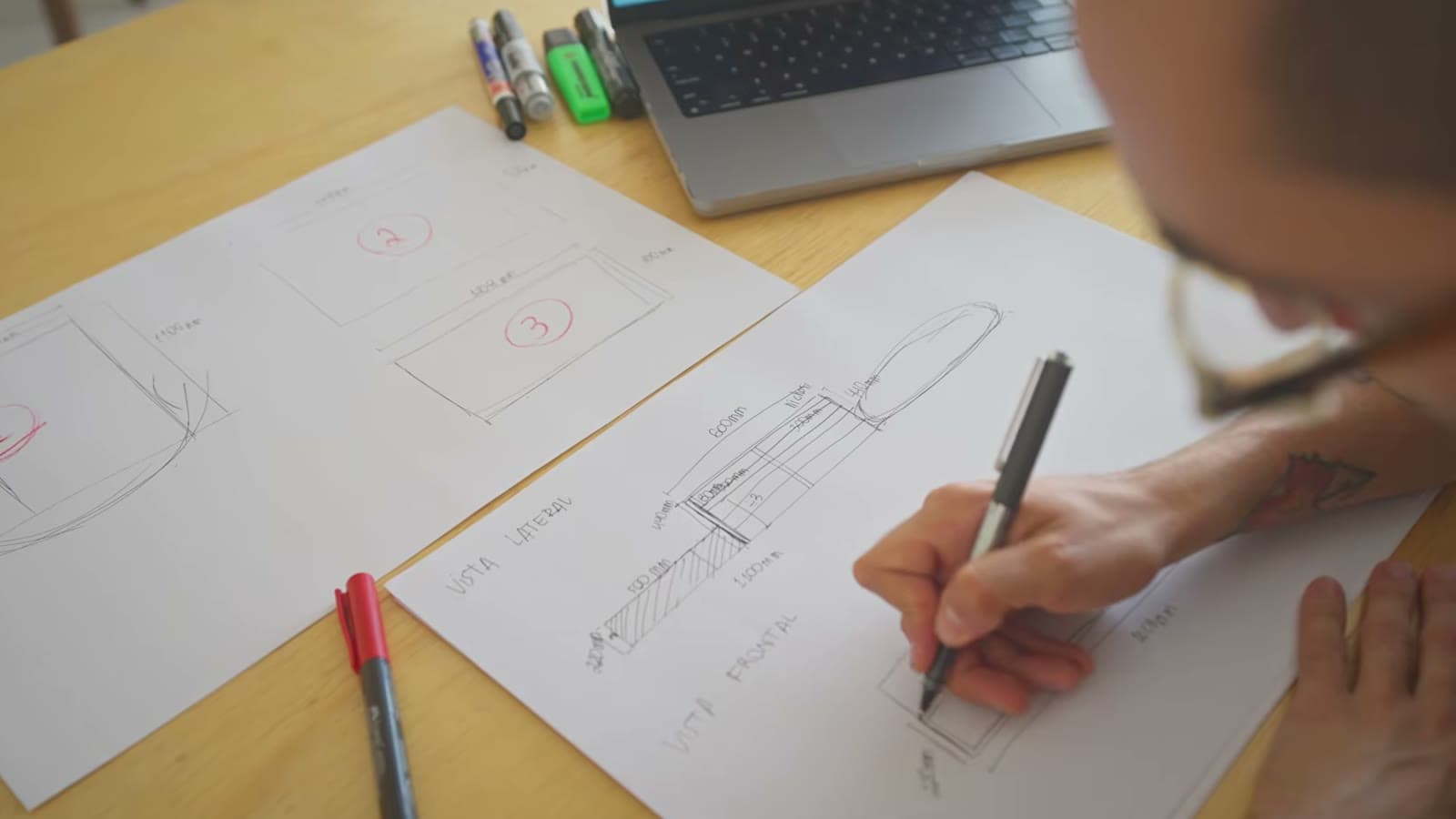 Mão desenhando com caneta o projeto da estrutura de madeira em folhas de papel, ao lado de um notebook e canetas coloridas