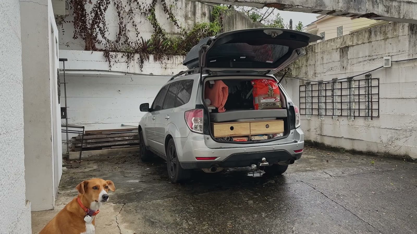 O Subaru estacionado num pátio com o porta-malas aberto e os gavetões de madeira à mostra, e a Nina sentada em primeiro plano