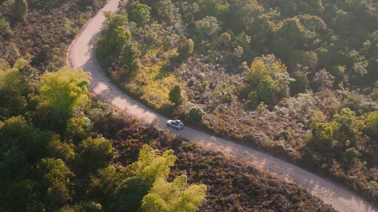 Vista aérea de uma estrada de chão fazendo curvas no meio da mata, com o carro pequeno lá embaixo