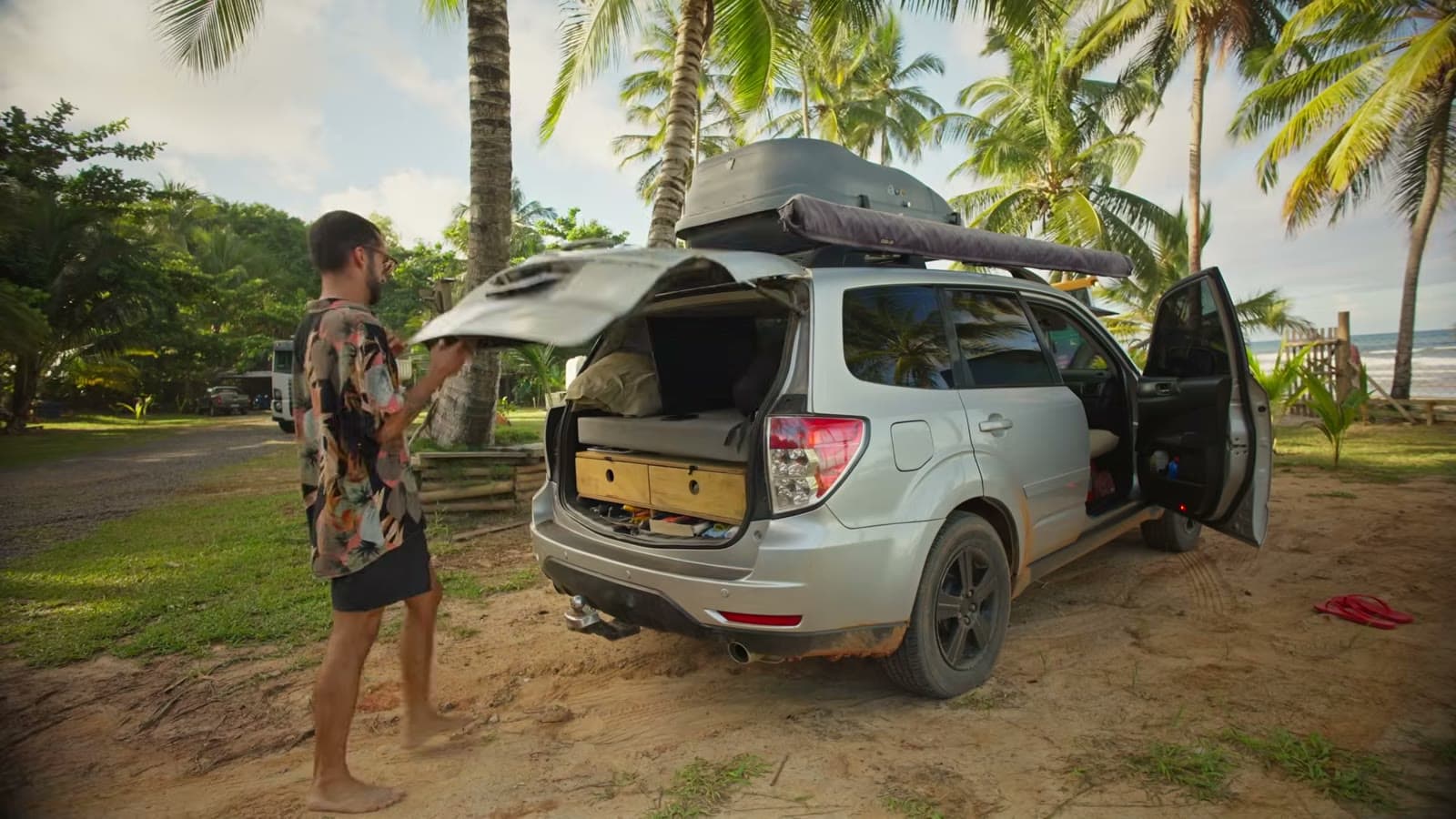 O Subaru estacionado na areia entre coqueiros, com o porta-malas e as portas abertos e os gavetões à mostra
