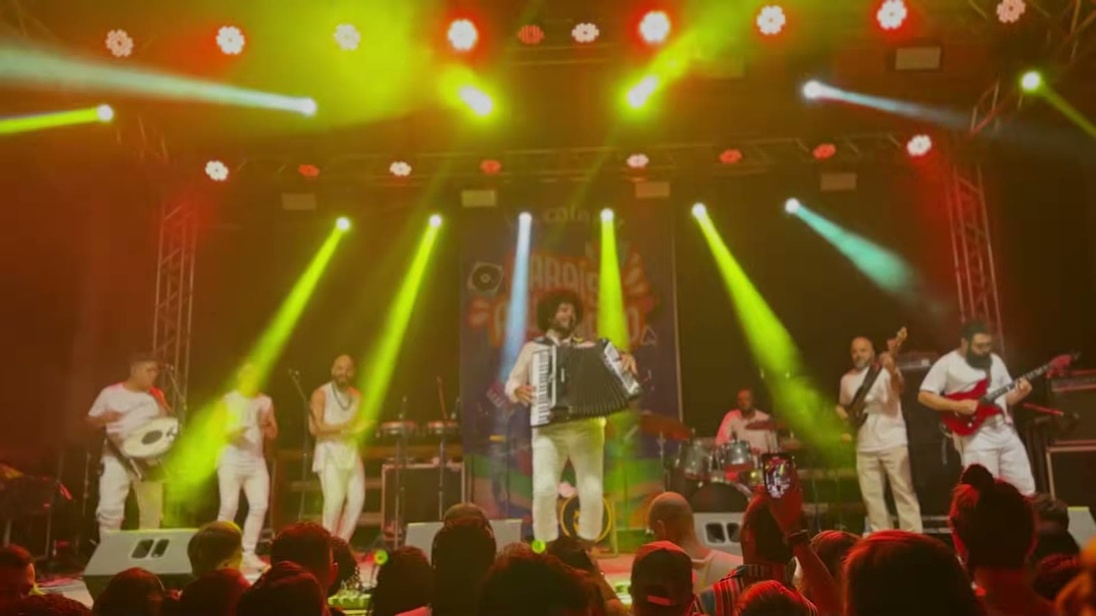 Banda de branco tocando sanfona e guitarra num palco com luzes verdes e vermelhas, diante do público