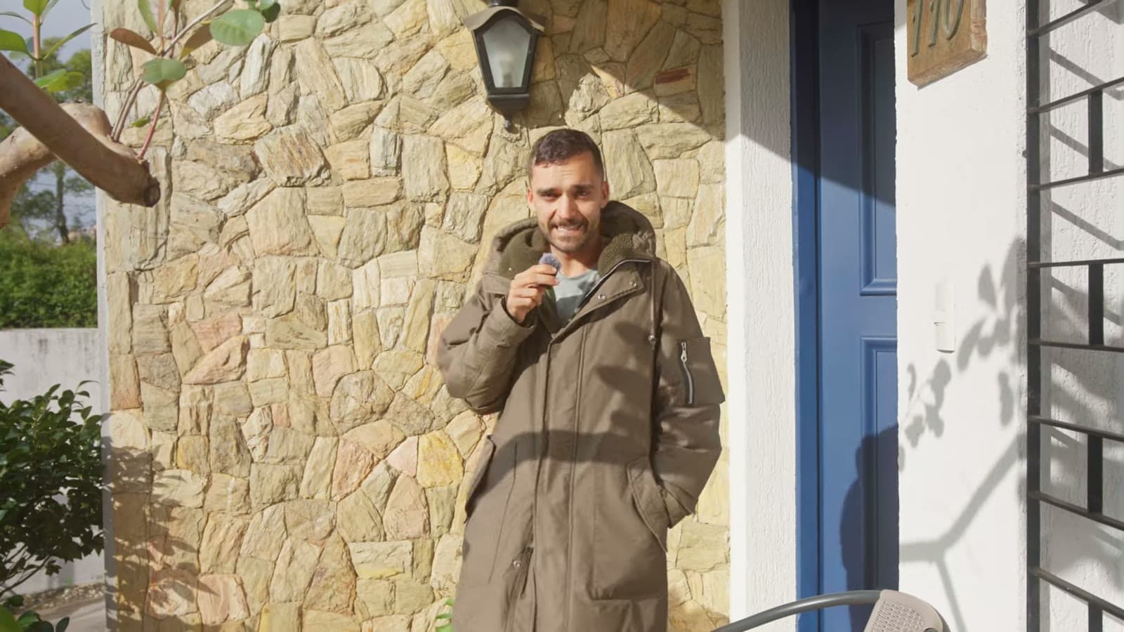 Nando in an olive-green jacket, holding the microphone, in front of a stone wall and a blue door