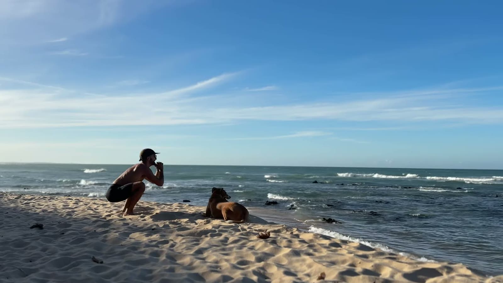 Nando crouching on the sand, facing the sea, with Nina lying beside him