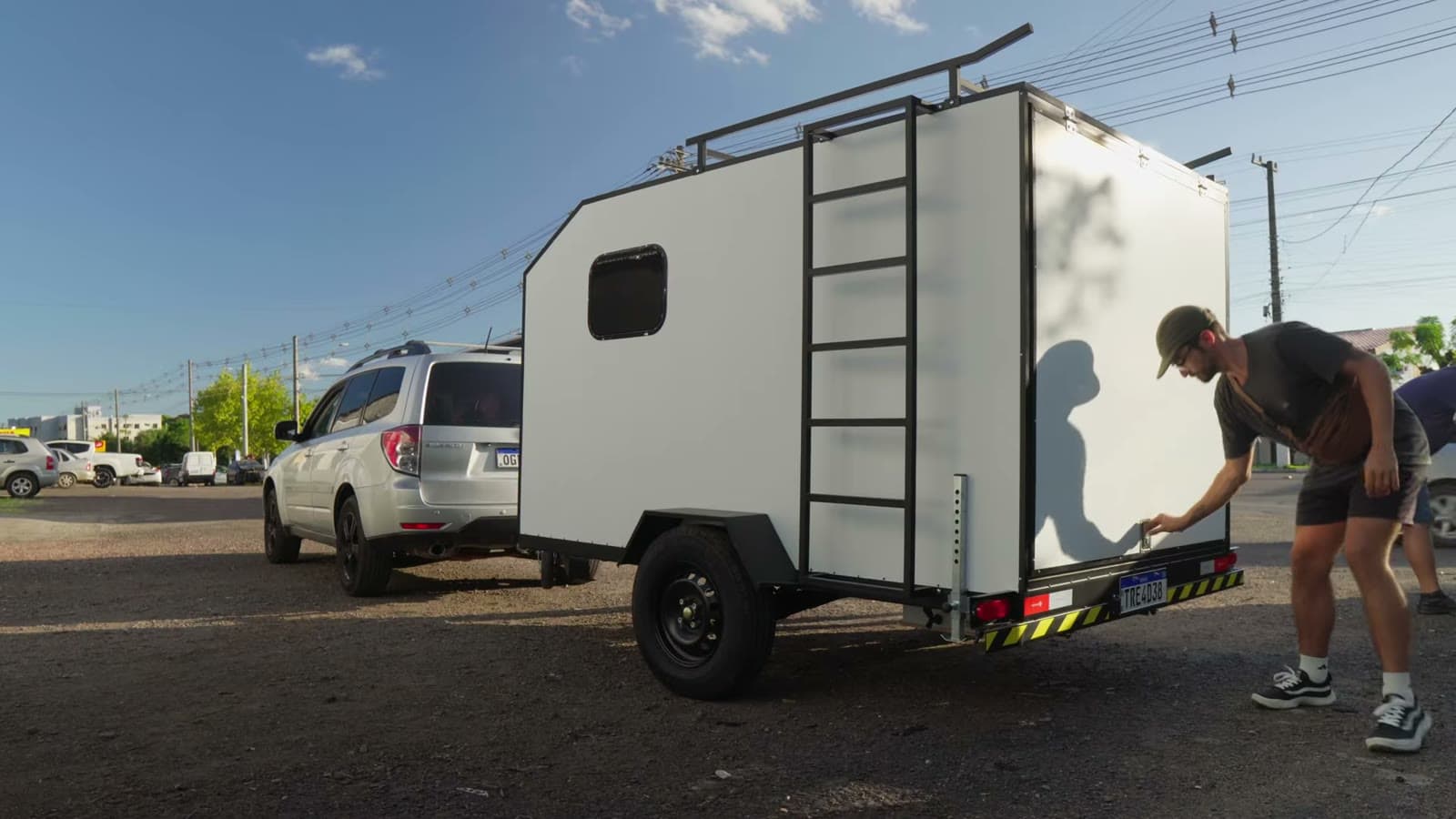 Nando adjusting the white mini trailer, with a ladder and roof rack, hitched to the car on a sunny street