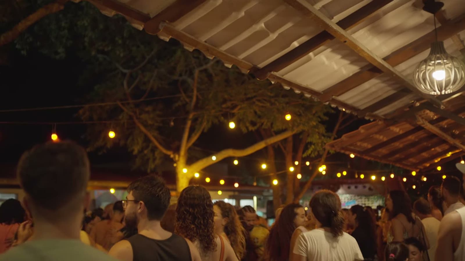 Crowd dancing forró under a wooden roof with string lights, at night