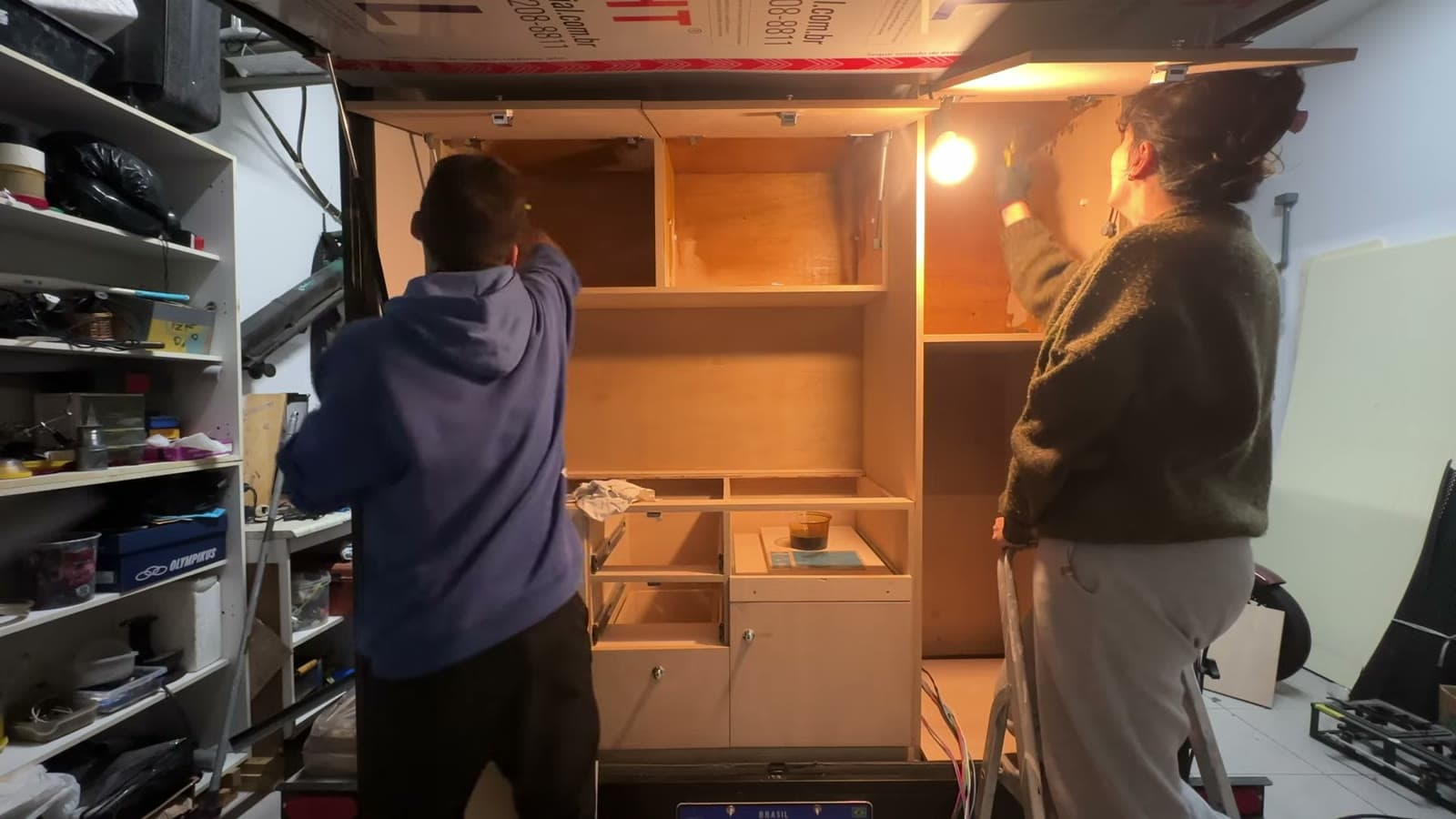 Two people building the wooden cabinets inside the mini trailer, lit by a light bulb