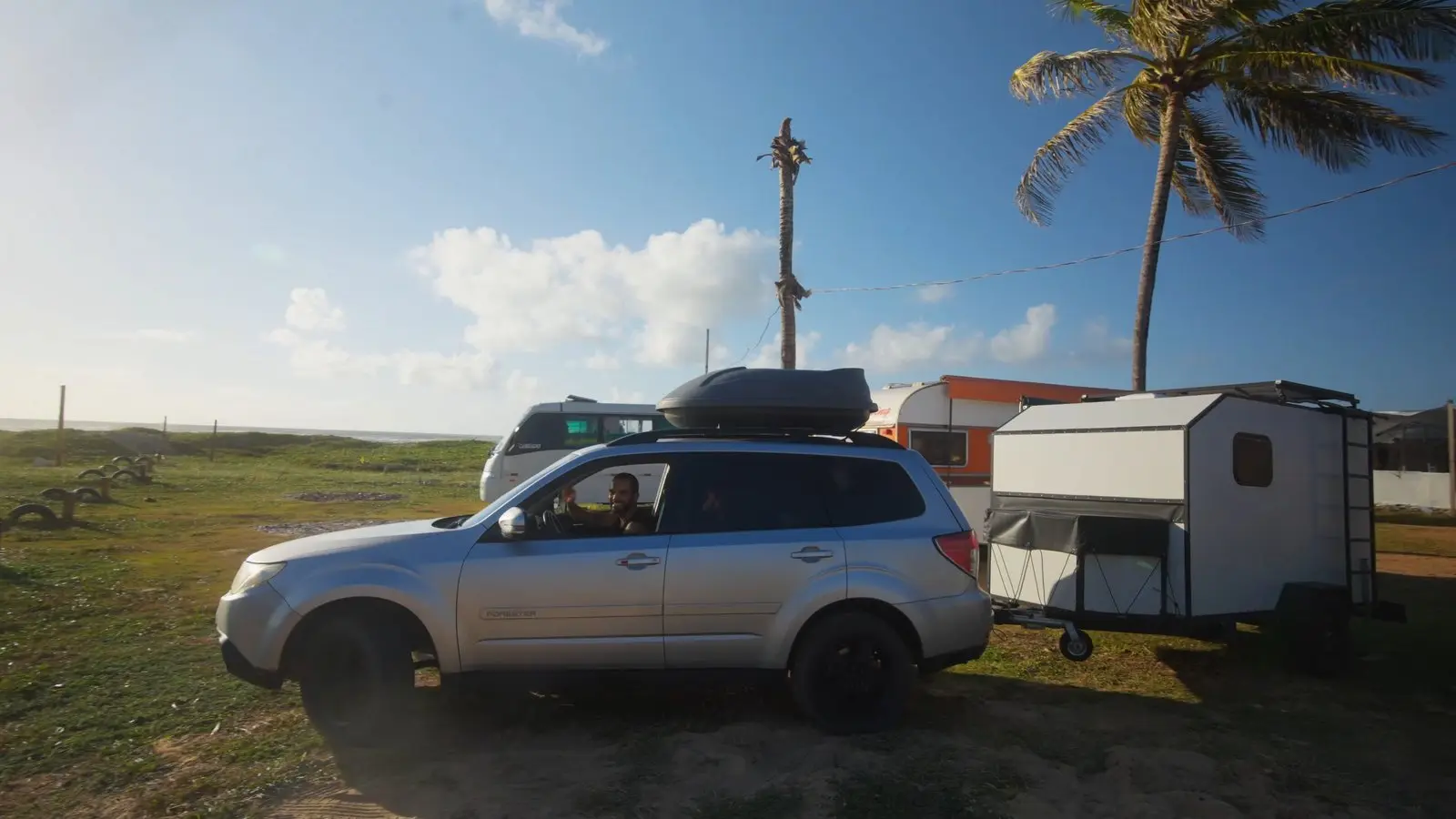 O carro prateado puxando o mini trailer branco num campo, com coqueiros e céu azul