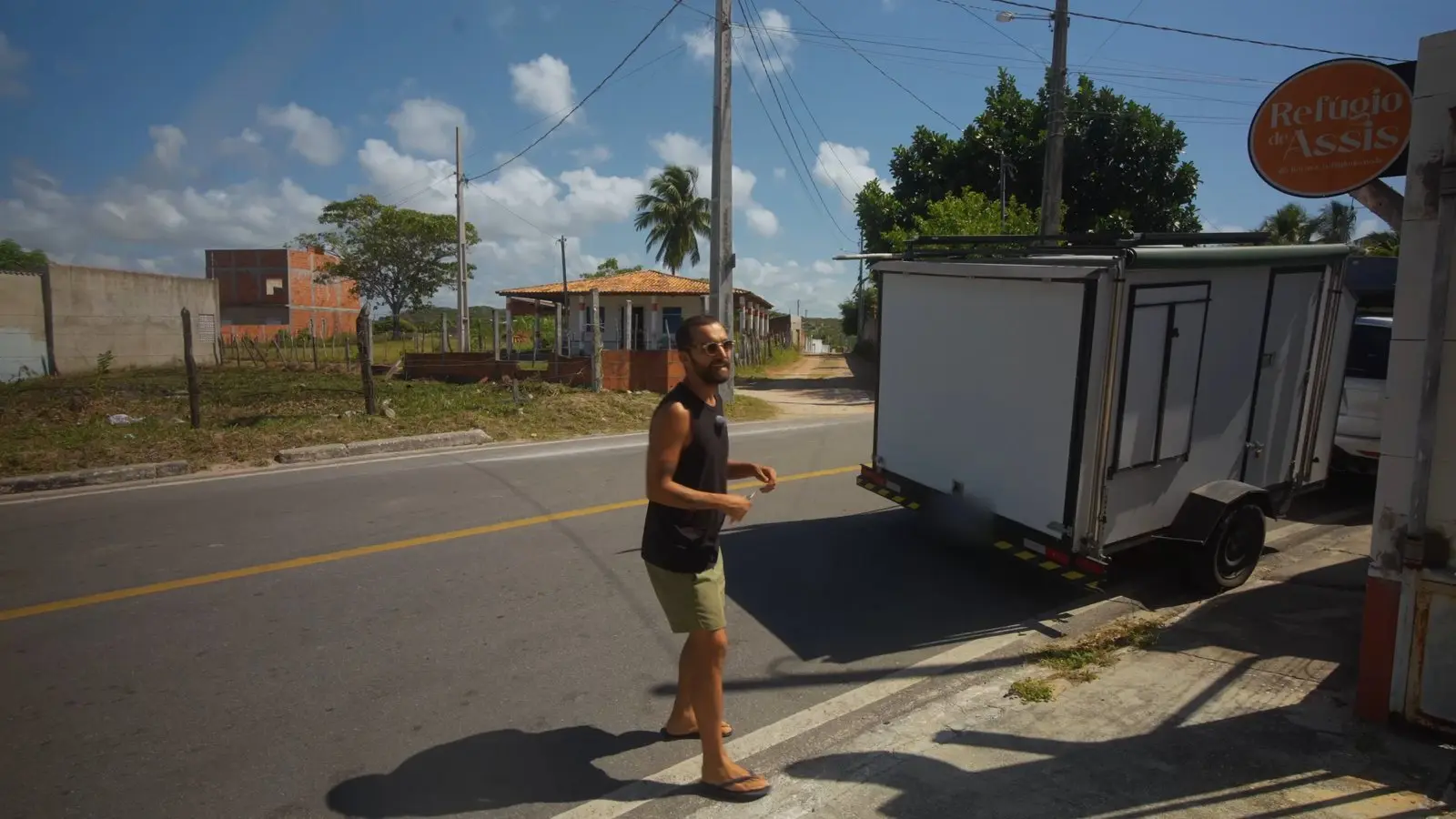 Nando na rua, na frente do portão da chácara, com o mini trailer e a placa da Refúgio de Assis ao fundo