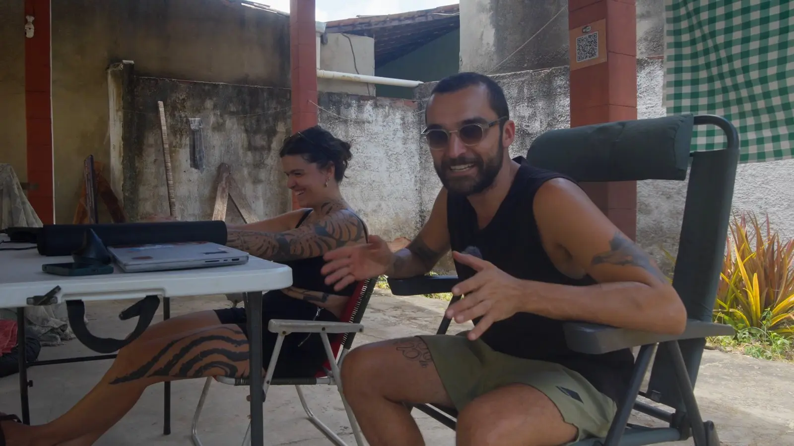 Nando e a Lelê sentados à mesa dobrável, com os computadores, rindo na chácara