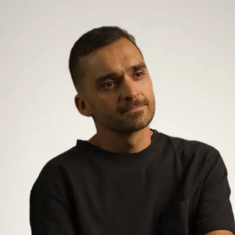 Nando Martins, retrato sentado, de camiseta preta, em fundo claro de estúdio